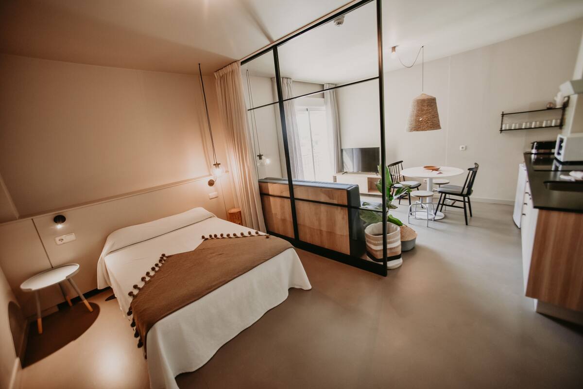 A modern hotel room with a bed, a small desk, a chair, a dining table, and a kitchen area.