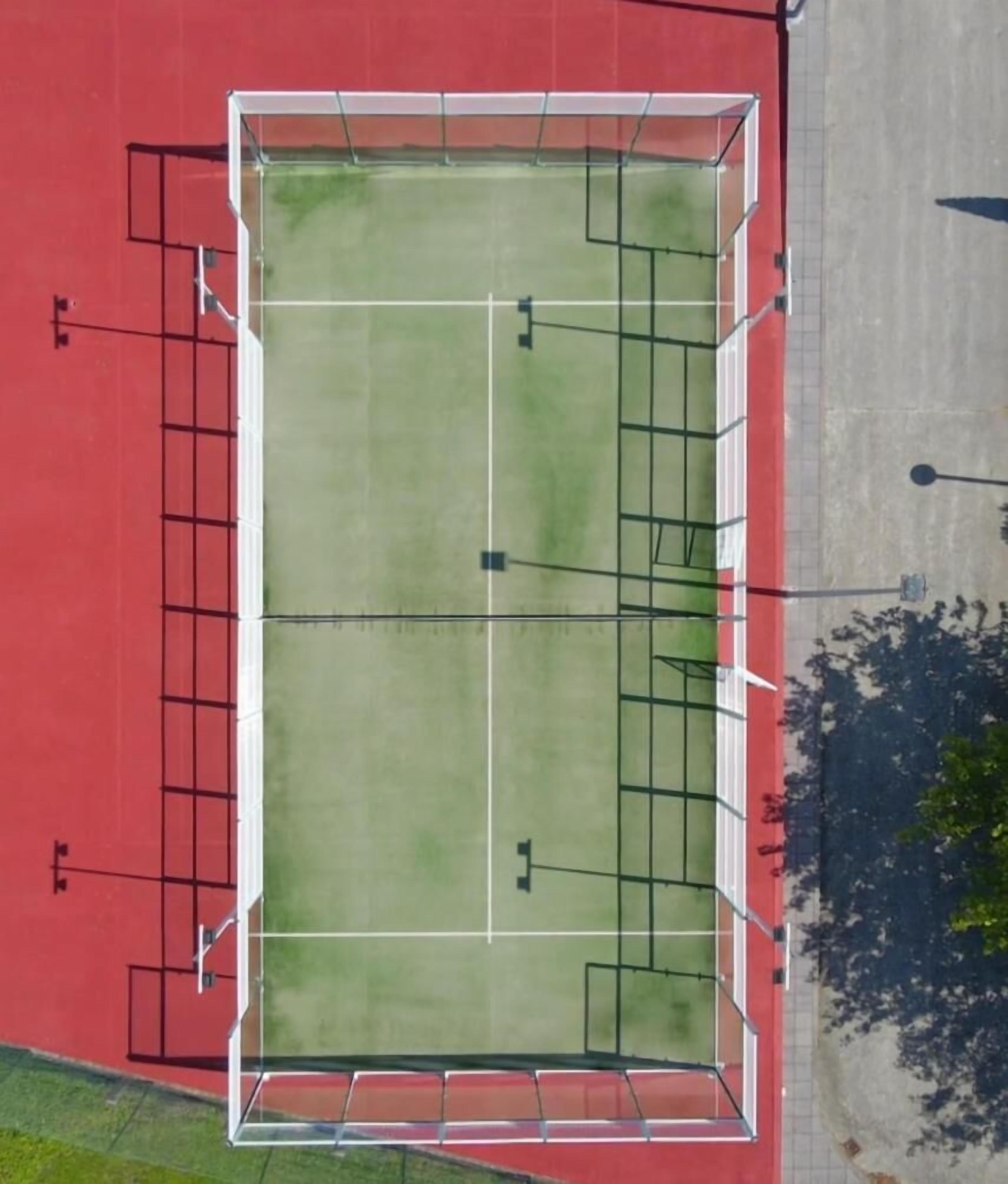 Vista aérea de un campo deportivo con líneas marcadas y porterías, rodeado por una pista roja y un área pavimentada con árboles.
