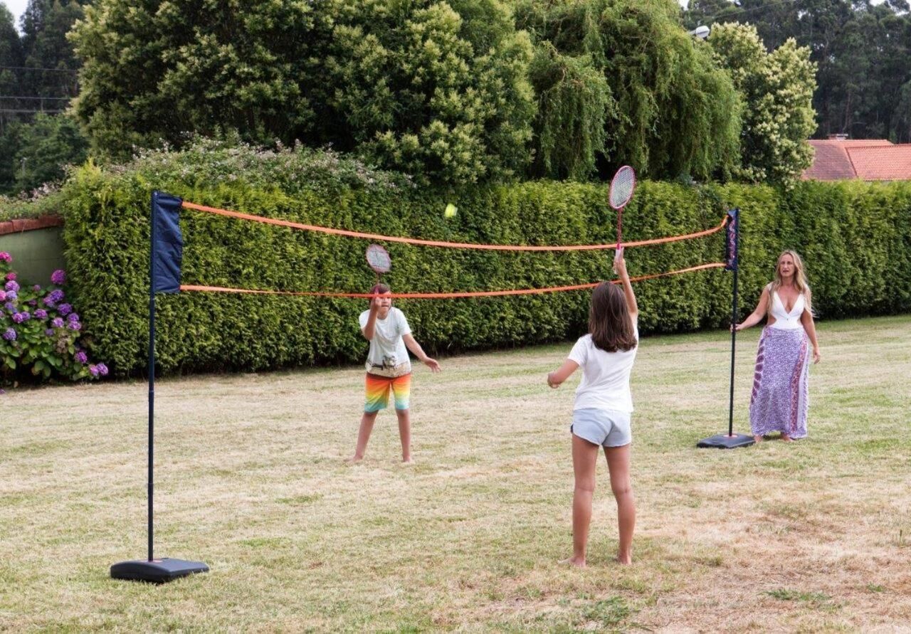Un partido de bádminton en un espacio al aire libre con césped, un seto y árboles de fondo.