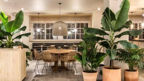 A modern dining area with a wooden table and wicker chairs, surrounded by large potted plants. - Apartamentos Dena (Meano)