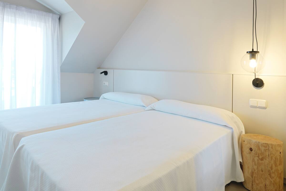 A bedroom with two beds, a bedside table, a nightstand, a lamp, and a window with curtains.