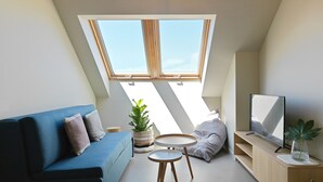 A modern loft bedroom with a skylight, a blue sofa, a small round table, a TV, and a potted plant. - Apartamentos Dena (Meano)