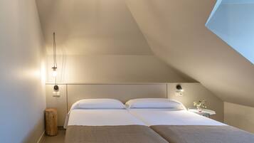 Un dormitorio moderno con techo inclinado, dos camas con ropa de cama a juego, lámparas de noche y un taburete de madera pequeño.