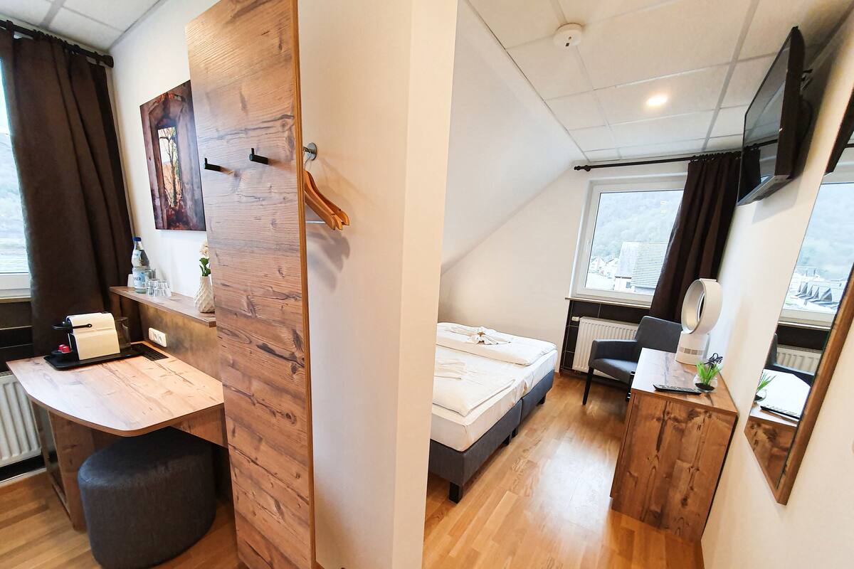 Quarto compacto com cama, escrivaninha, cadeira, radiador e janela com vista.