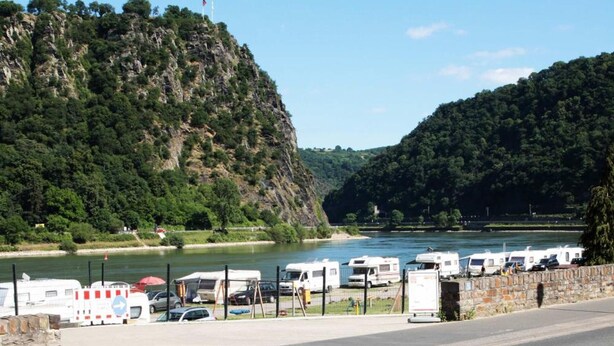A view from a deck overlooking a river with RVs parked along the shore, a mountain in the background, and a wooden table with a mat and a number sign. - Hotel Winzerhaus Gaertner an der Loreley (Sankt Goar)
