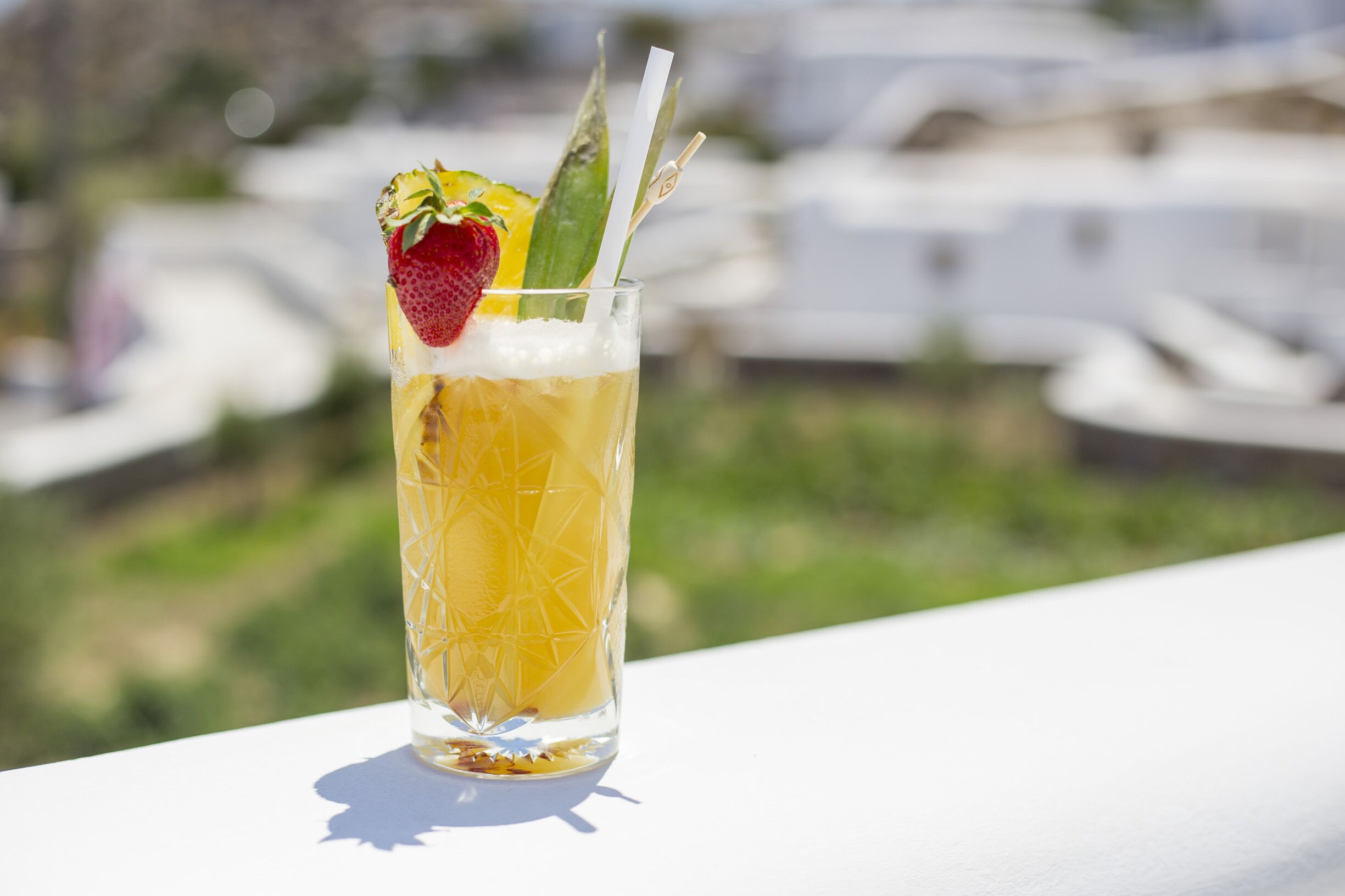 A tall glass with a straw, containing a yellow beverage, garnished with a strawberry and a slice of lime, placed on a white surface with a blurred outdoor background.