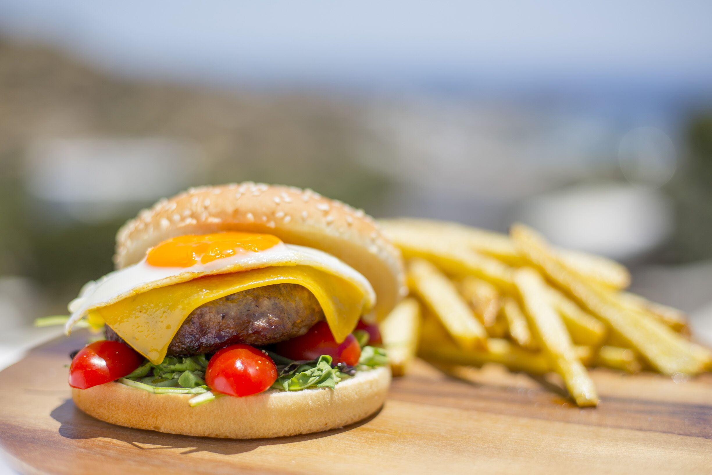 A hamburger with a fried egg, cheese, lettuce, and tomatoes, accompanied by a side of French fries on a wooden board.