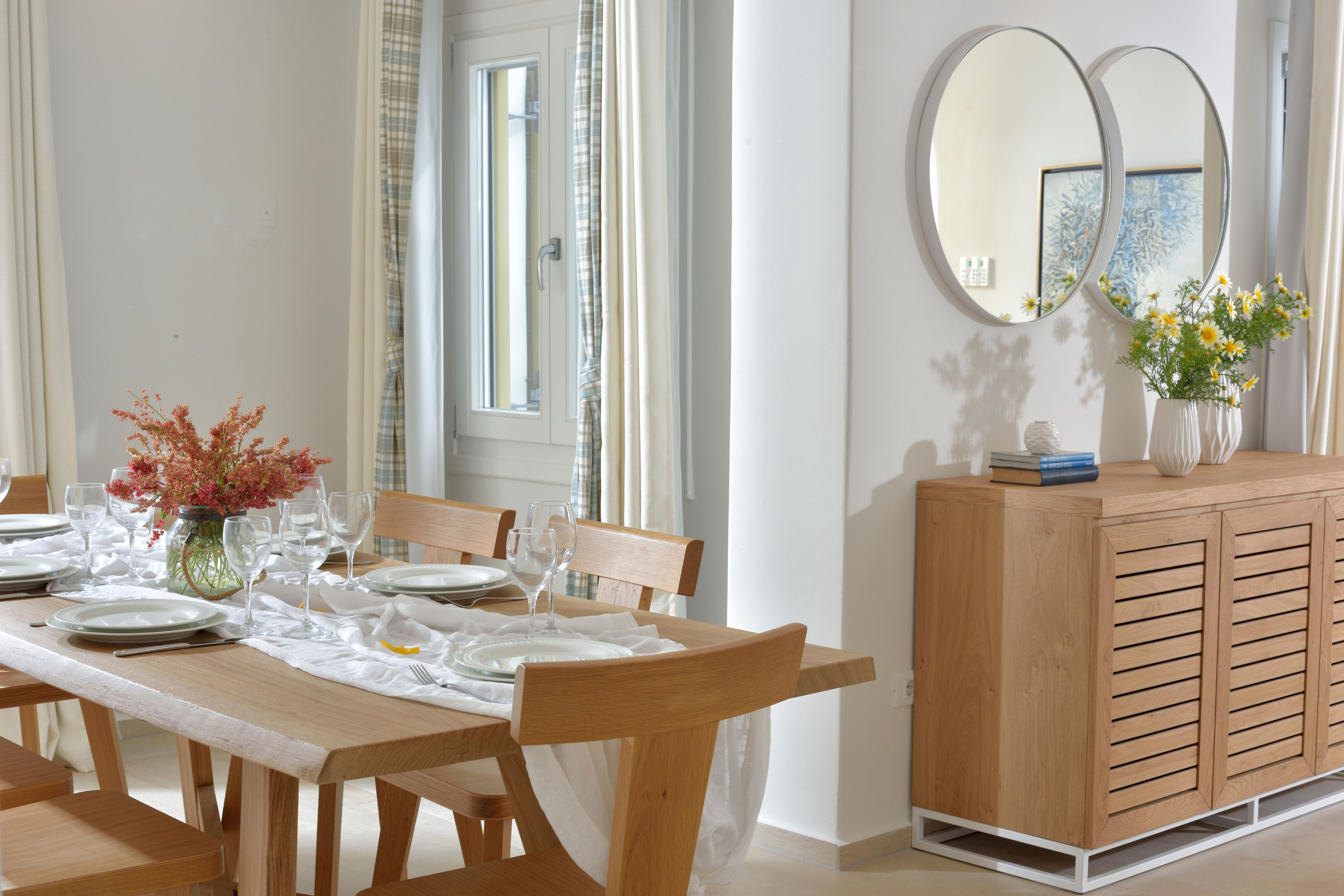 A dining area with a wooden table set for six, a wooden cabinet with a vase of flowers, and two round mirrors on the wall.