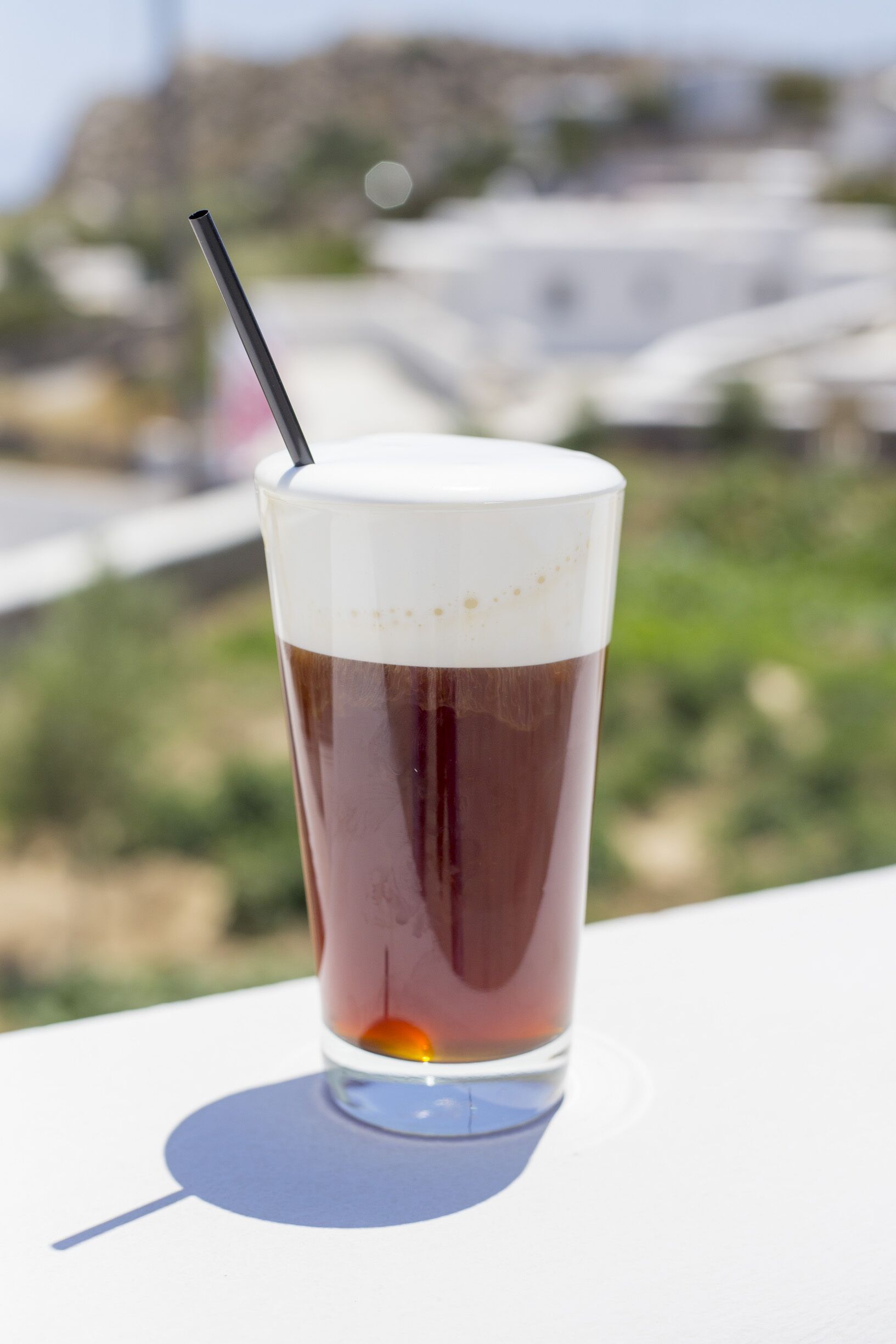 Un vaso alto de café helado con una pajita, sobre una superficie blanca con un fondo desenfocado al aire libre.