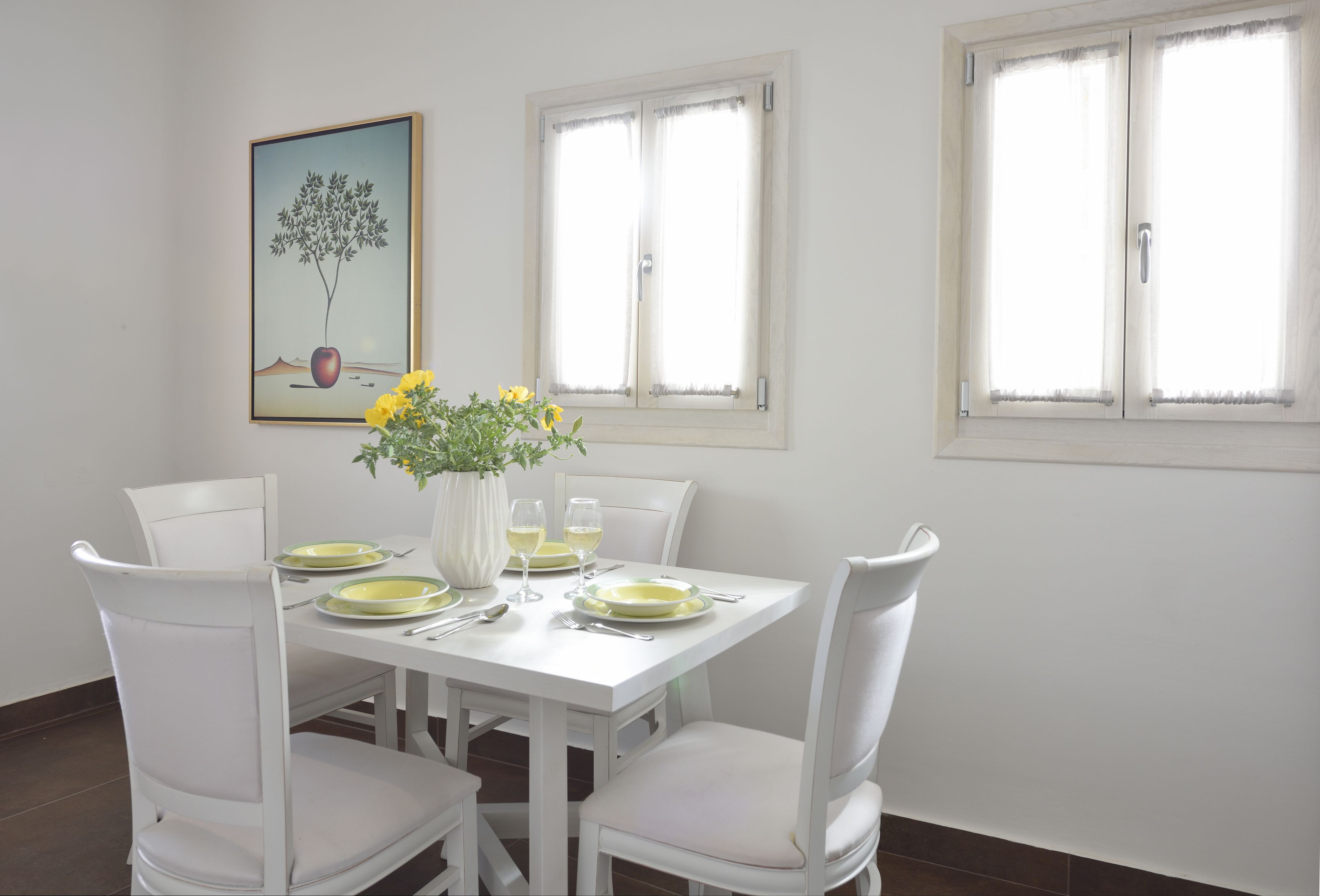 Un coin repas avec une table blanche dressée pour quatre personnes, des chaises blanches, un vase rempli de fleurs jaunes et un tableau accroché au mur.