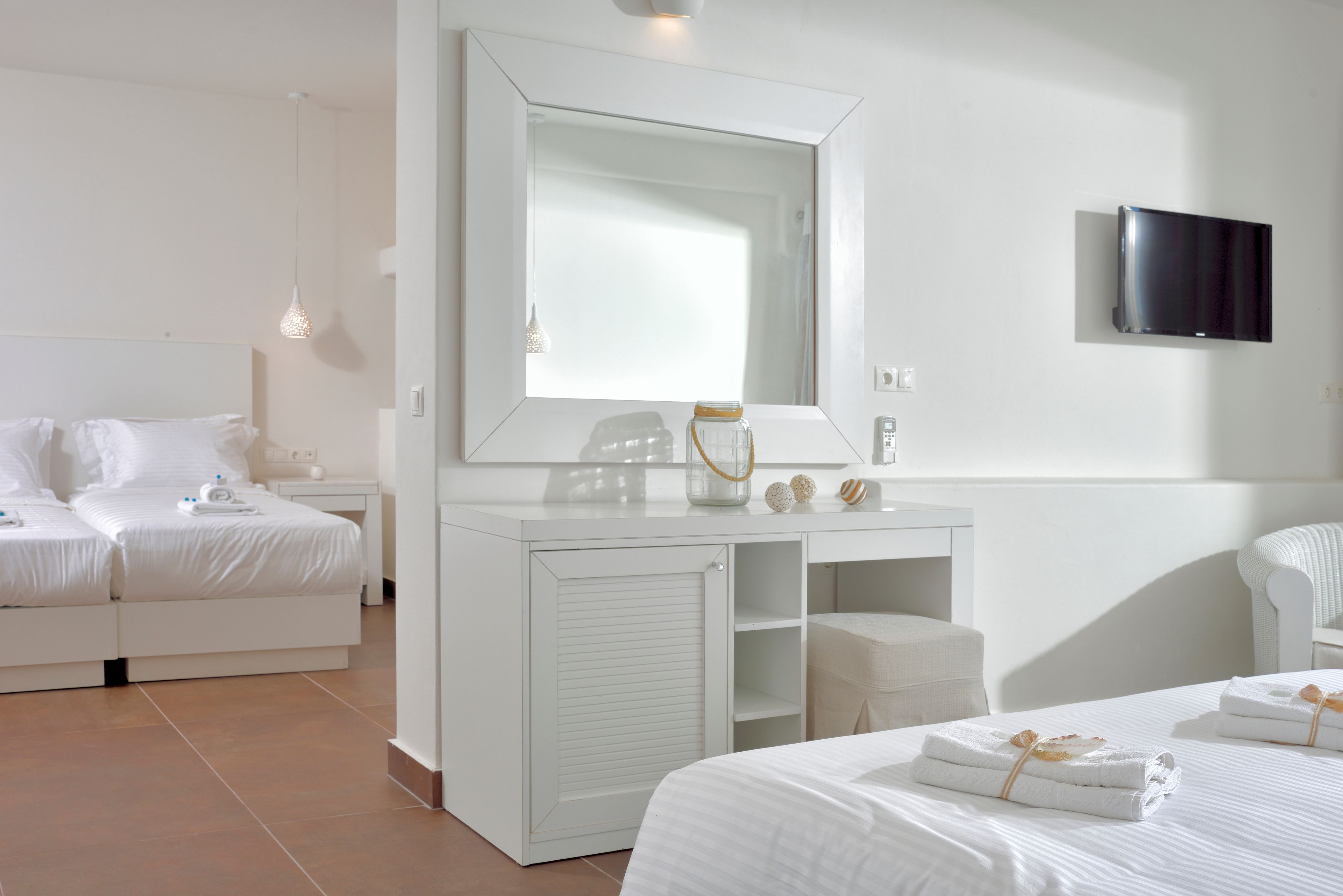 A hotel room with a large mirror, a flat-screen TV, a bed with white linens, a bedside table, and a small seating area.