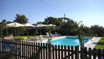 A swimming pool with lounge chairs and umbrellas, surrounded by a wooden fence and greenery.