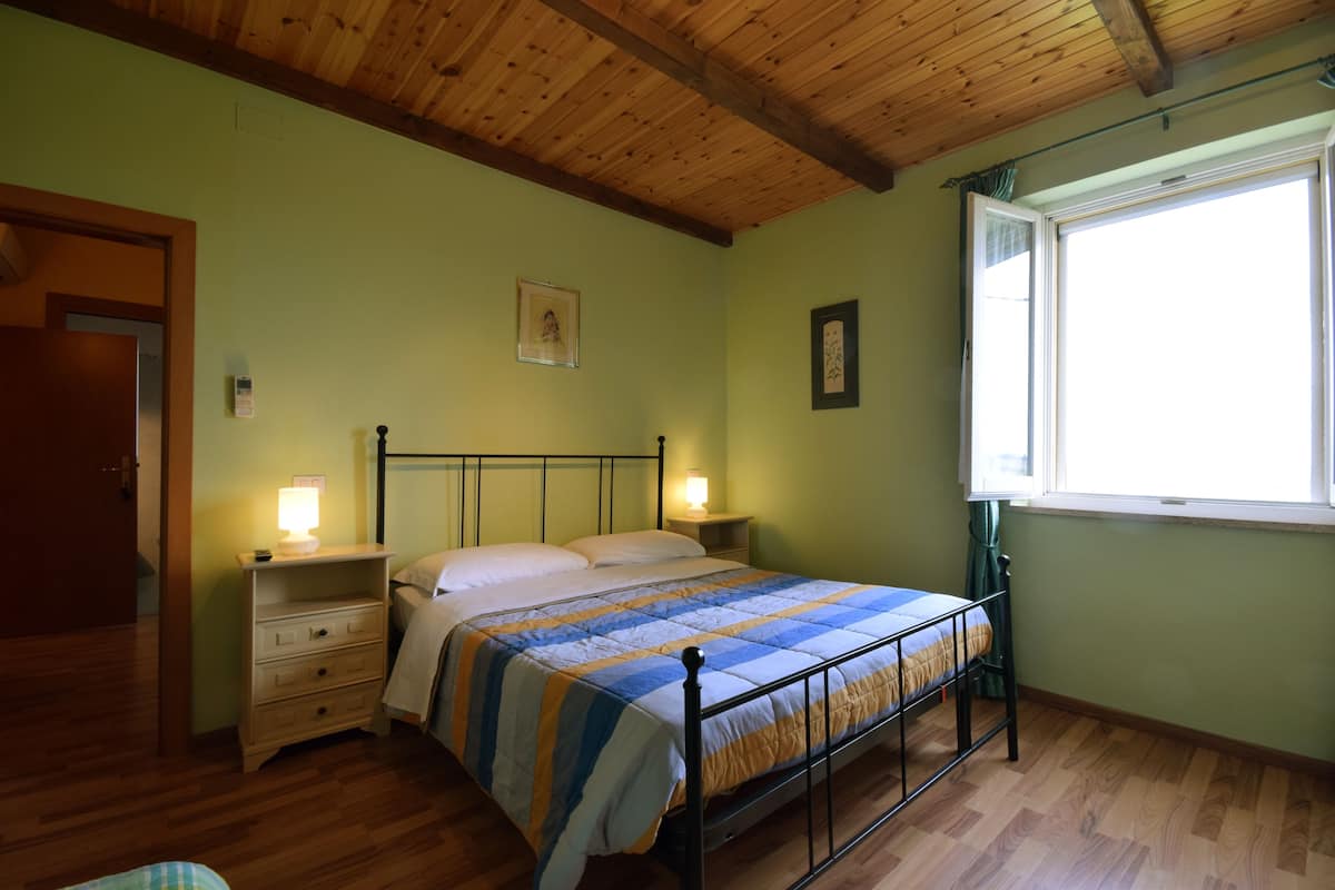 A bedroom with a wooden ceiling, a bed with a striped bedspread, bedside lamps, a nightstand, and a window with curtains.