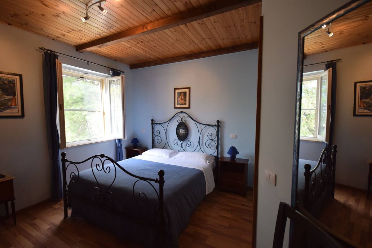 A bedroom with a wooden ceiling, a bed with a metal frame, a nightstand with a lamp, and a window with curtains.