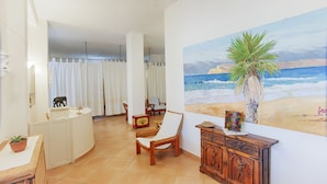 Quarto com um mural de praia, uma cadeira de madeira, uma pequena mesa com uma planta e um armário com objetos decorativos.
