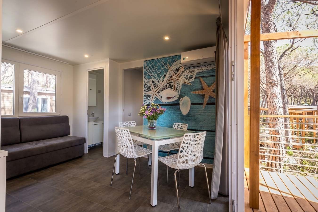 Zona pranzo con tavolo in vetro e sedie bianche, divano grigio e parete decorata con conchiglie e stelle marine.