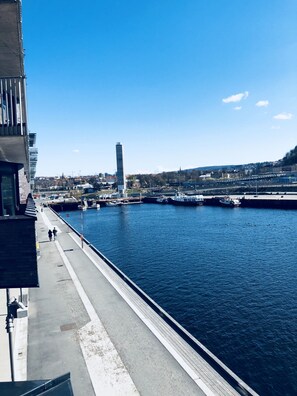 A waterfront promenade with a clear view of the water, a tall monument, and a distant cityscape.
