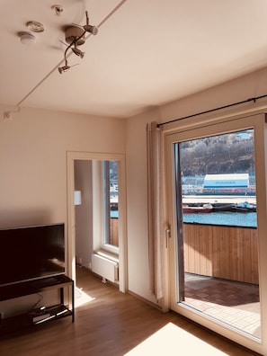 Ett rum med en tv på ett stativ, en skjutdörr i glas som leder ut till ett utomhusområde med utsikt över ett fartyg och en byggnad med skylten "SKANSKA".