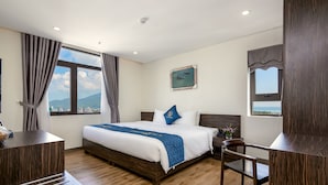 A hotel room with a large bed, a desk, and a chair. There is a window with a view of the city and mountains. - Sepon Blue Hotel (Da Nang)
