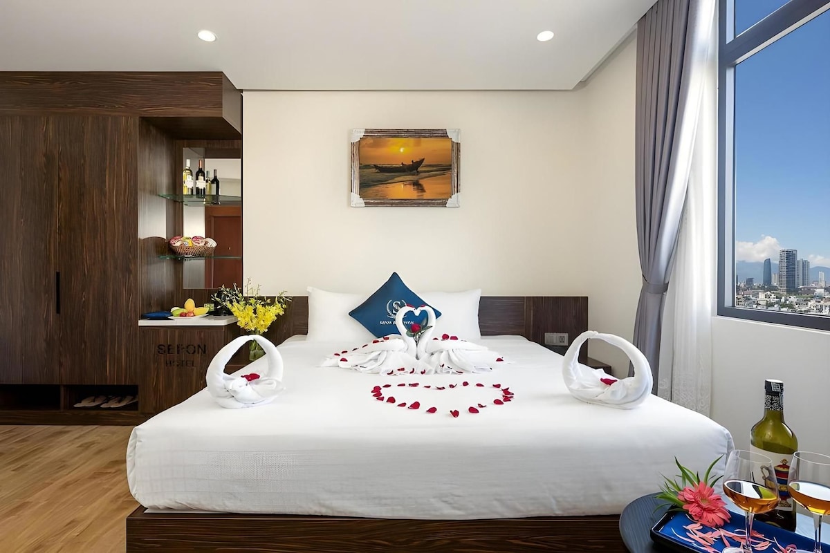 A hotel room with a bed, a city view, a painting, and a minibar.