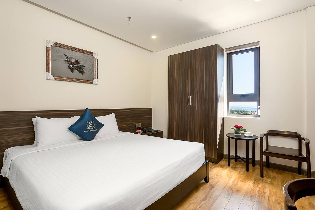 A hotel room with a large bed, a wooden wardrobe, a small table with a vase of flowers, and a window with a view.