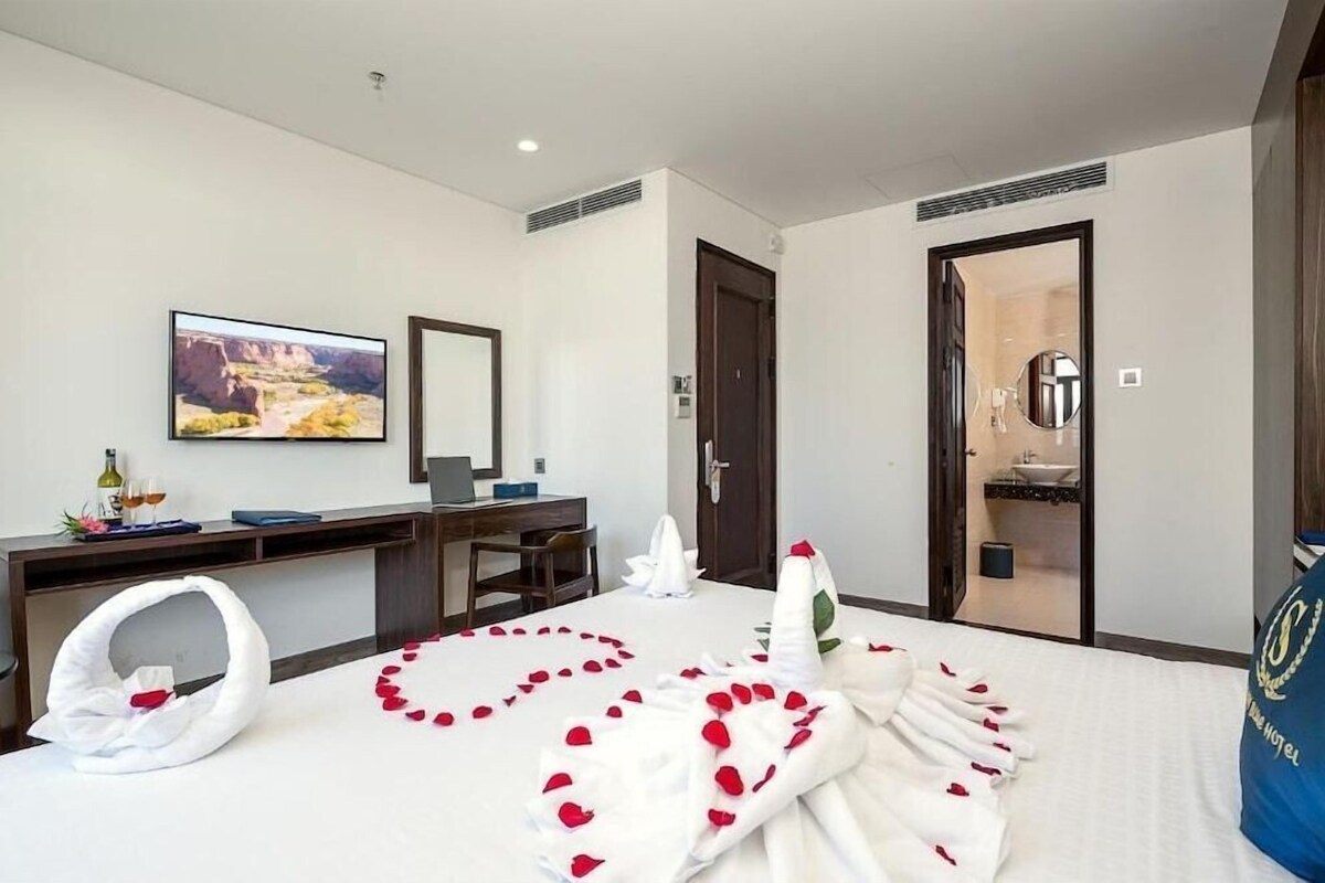 A hotel room with a bed, a desk, a chair, a mirror, a painting, and a bathroom.