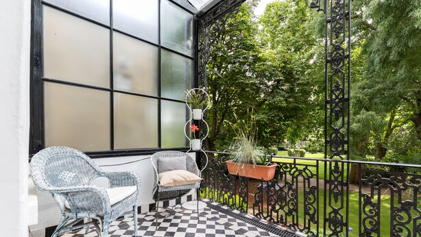 A balcony with a checkered floor, a wicker chair, a potted plant, and a view of a green garden. - Philbeach Balcony- 2bed flat in Earls Ct (London)