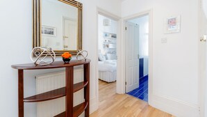 A hallway with a wooden console table, a mirror, and a radiator. There is a door leading to a bedroom with a white bed and a blue-tiled floor. - Philbeach Balcony- 2bed flat in Earls Ct (London)