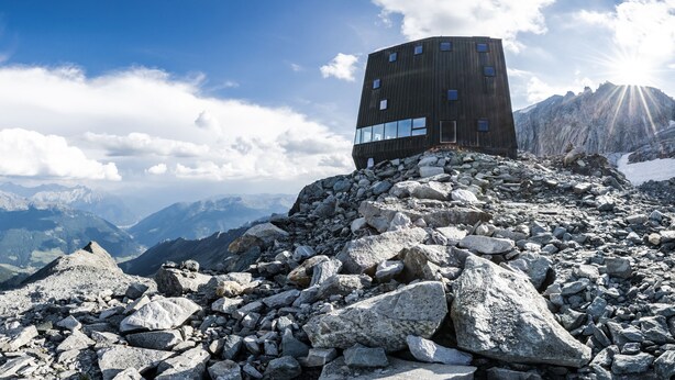 Um edifício moderno, com vários andares e grandes janelas, localizado no topo de uma montanha rochosa, com vista panorâmica para montanhas distantes e um céu claro.