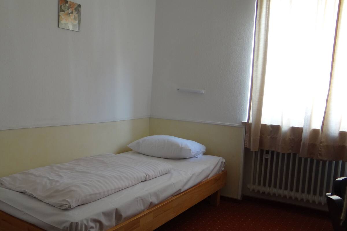 A small, single-bed room with a wooden frame, white bedding, and a radiator near the window.