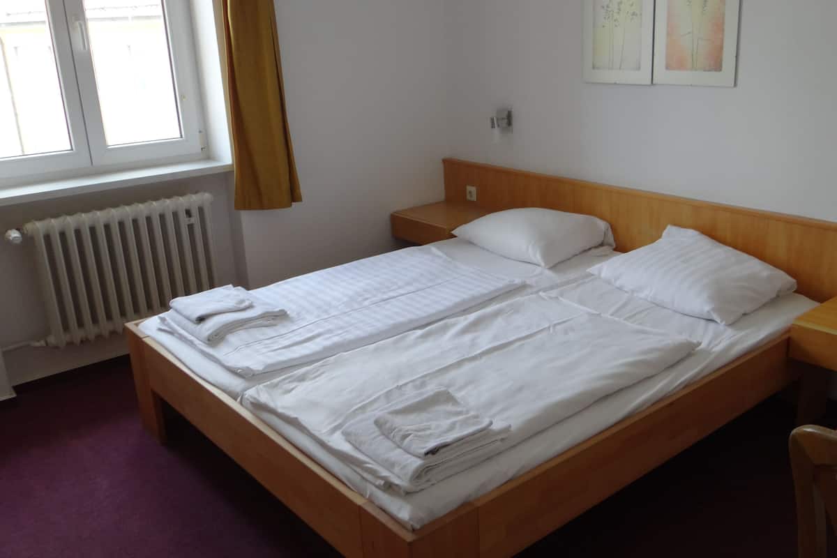 A single bed with white linens, a wooden headboard, a nightstand, a red table, and a window with curtains.