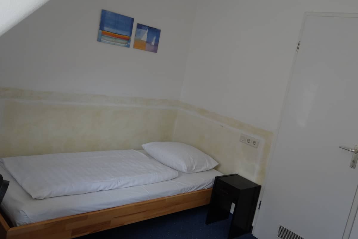 A small, simple room with a single bed, a door, and a wall-mounted light switch.