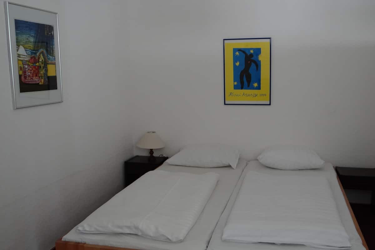 A twin bed with white bedding in a room with white walls. There is a framed artwork above the bed and a small table with a lamp beside it.
