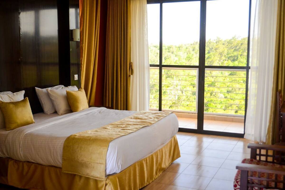 A hotel room with a large bed, a balcony with a view of greenery, and a wooden chair.