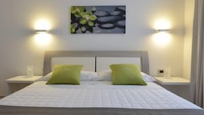 A neatly arranged bedroom with a bed, two green pillows, two bedside tables, and a wall-mounted light fixture. - White (Porto Cesareo)