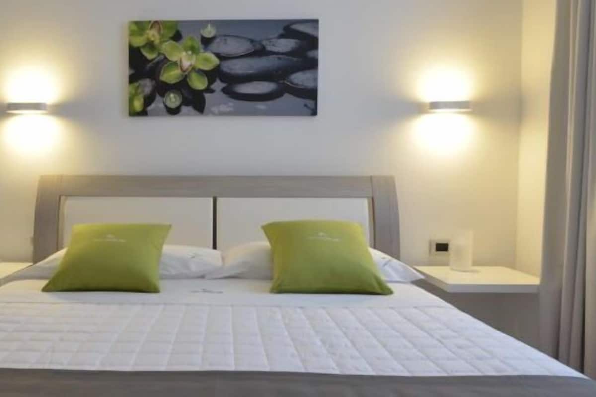 A neatly arranged bedroom with a bed, two green pillows, two bedside tables, and a wall-mounted light fixture.
