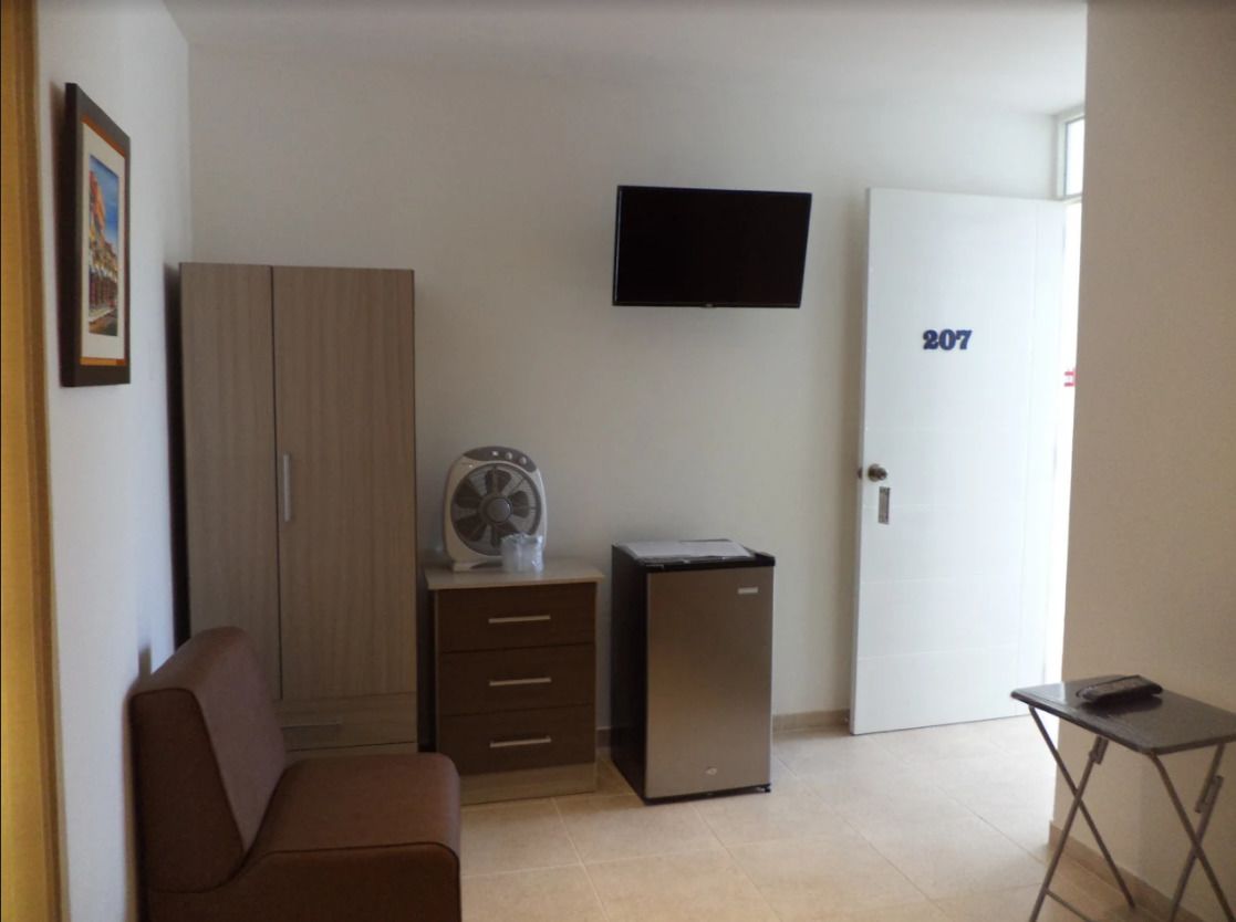 Una habitación con un sofá marrón, una mesita, un armario, un refrigerador, un ventilador y un televisor montado en la pared.