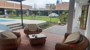 Een overdekt terras met rieten meubels en een kleine tafel, een zwembad en uitzicht op een woonwijk met een auto en gebouwen.