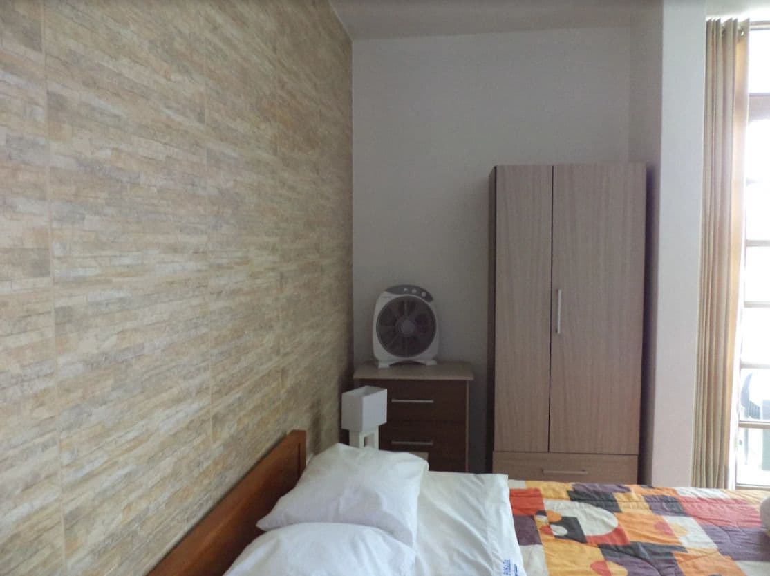 Ein Schlafzimmer mit einer Wand im Backstein-Look, einem Holzschrank, einem Bett mit Patchworkdecke und einem weißen Ventilator auf einem Nachttisch.