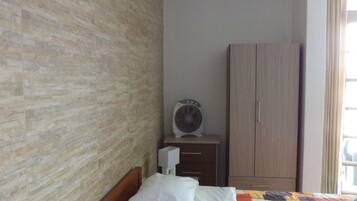 Ein Schlafzimmer mit einer Wand im Backstein-Look, einem Holzschrank, einem Bett mit Patchworkdecke und einem weißen Ventilator auf einem Nachttisch.
