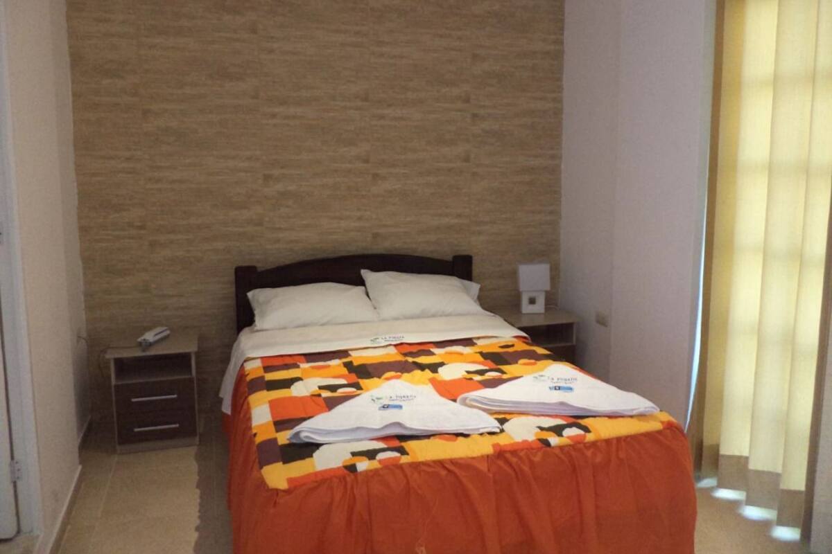A neatly made bed with a patterned bedspread, a bedside table with a lamp, and a window with curtains.