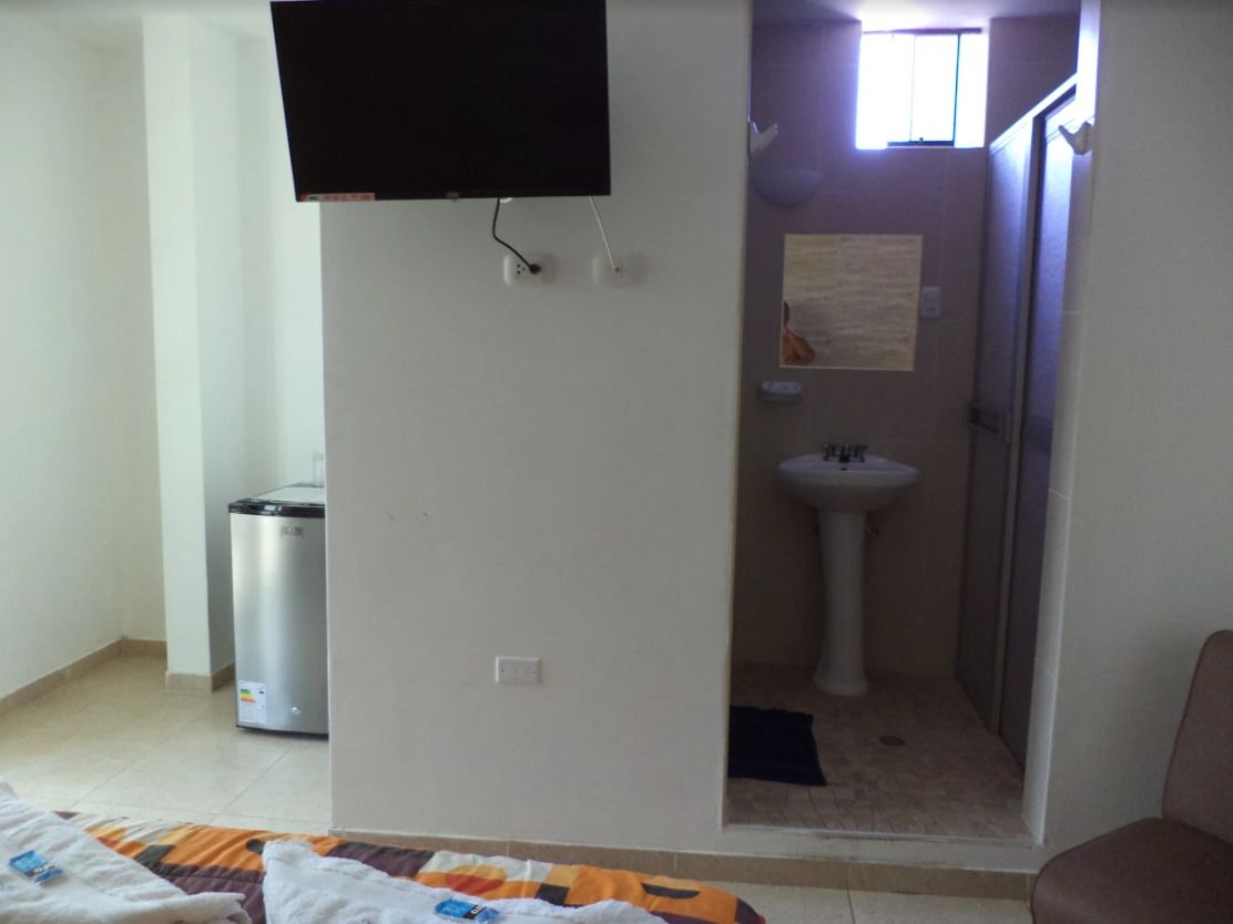 Une chambre avec un lit, un téléviseur fixé au mur, une petite poubelle et une salle de bain visible à travers une porte ouverte.