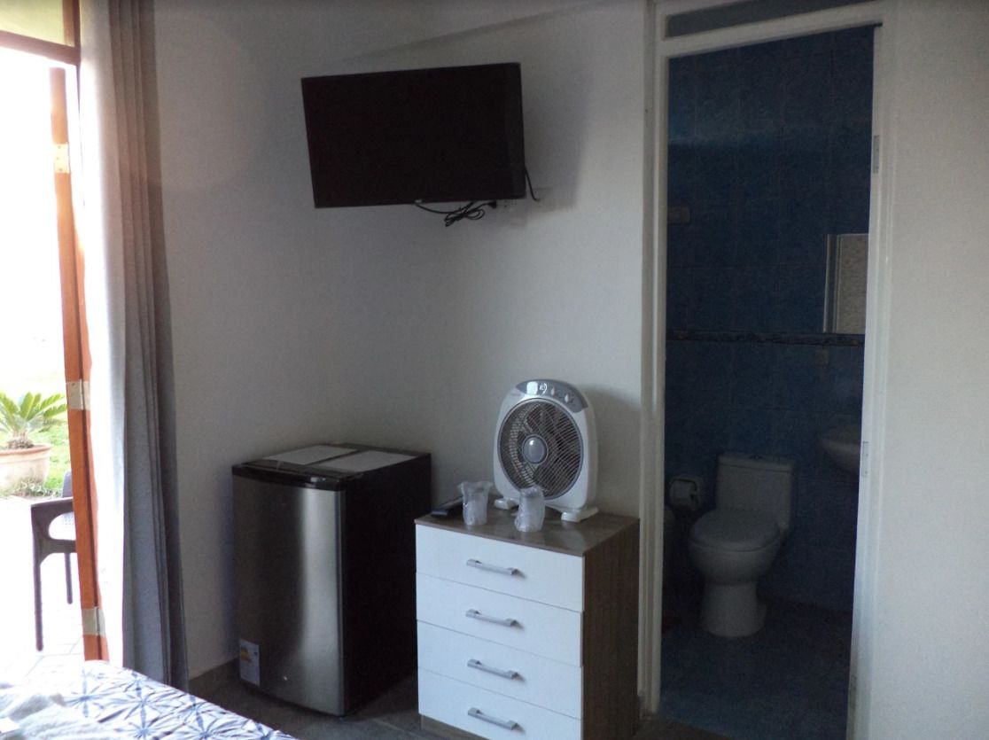 A compact room with a bed, a small refrigerator, a fan, a television, and a bathroom with a toilet.