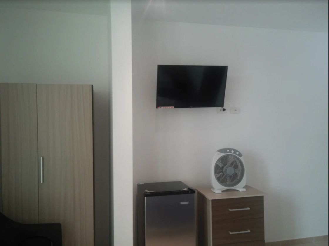 Una habitación con televisión montada en la pared, un refrigerador pequeño y un ventilador sobre un mueble de madera.