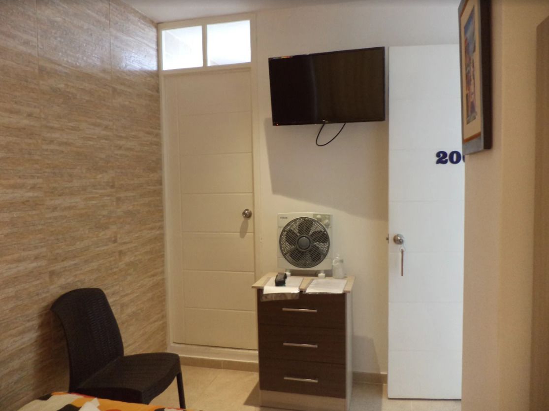 Una habitación con cama, una silla, cómoda, televisor montado en la pared y una puerta con un número.