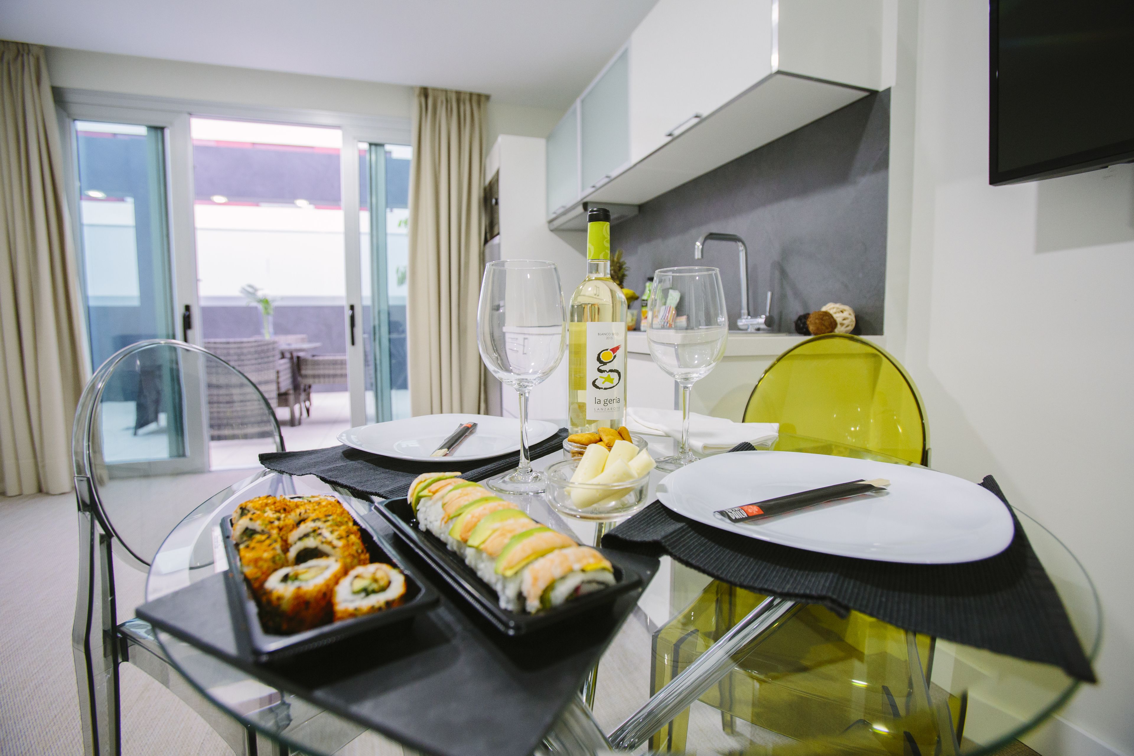 Comedor con una mesa de cristal dispuesta para dos personas, que incluye sushi, una botella de vino blanco y un bol de fruta.