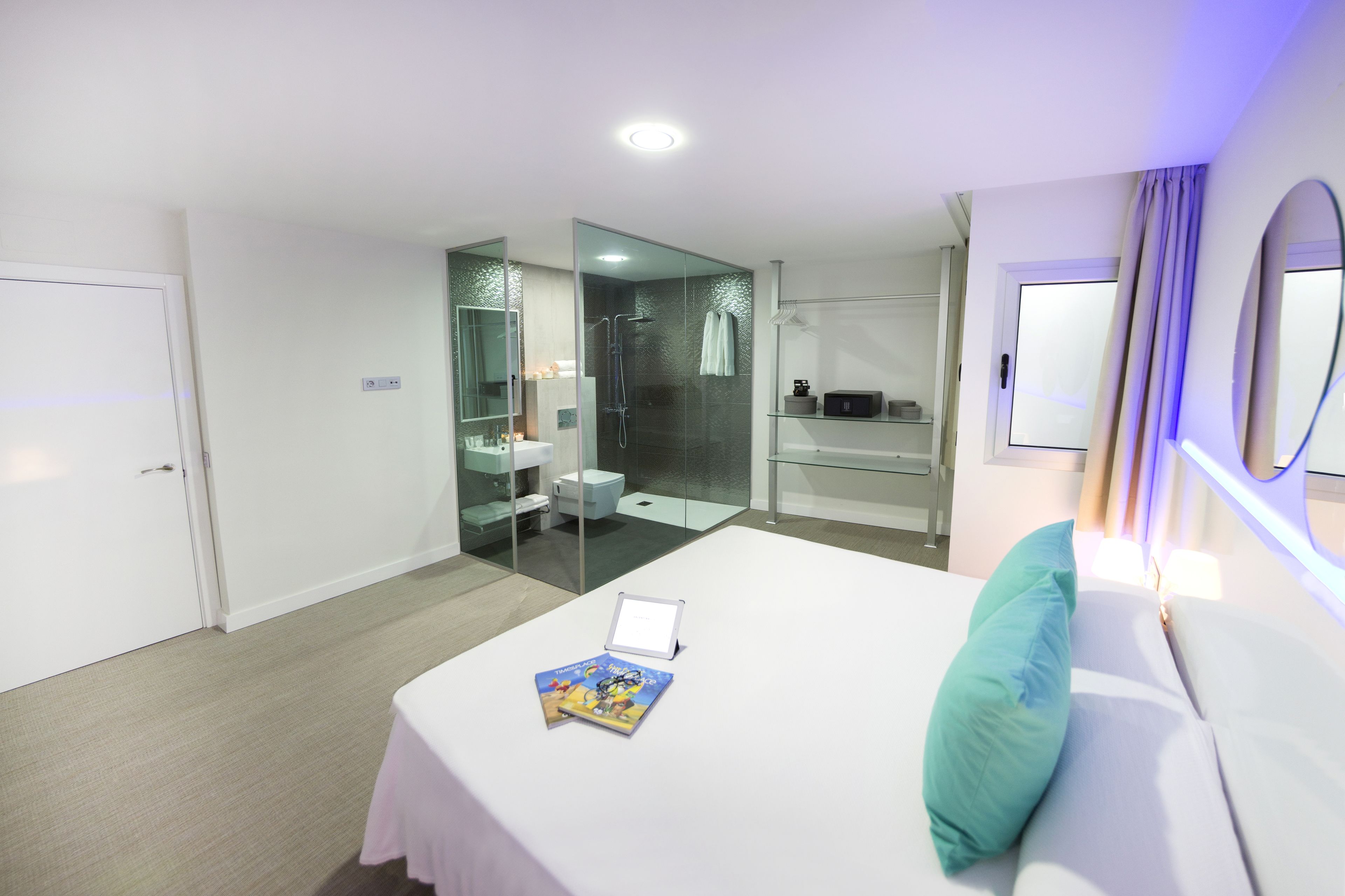 Una habitación de hotel moderna con cama, mesita de noche con un libro, zona de ducha con mampara de vidrio y baño con lavamanos y espejo.