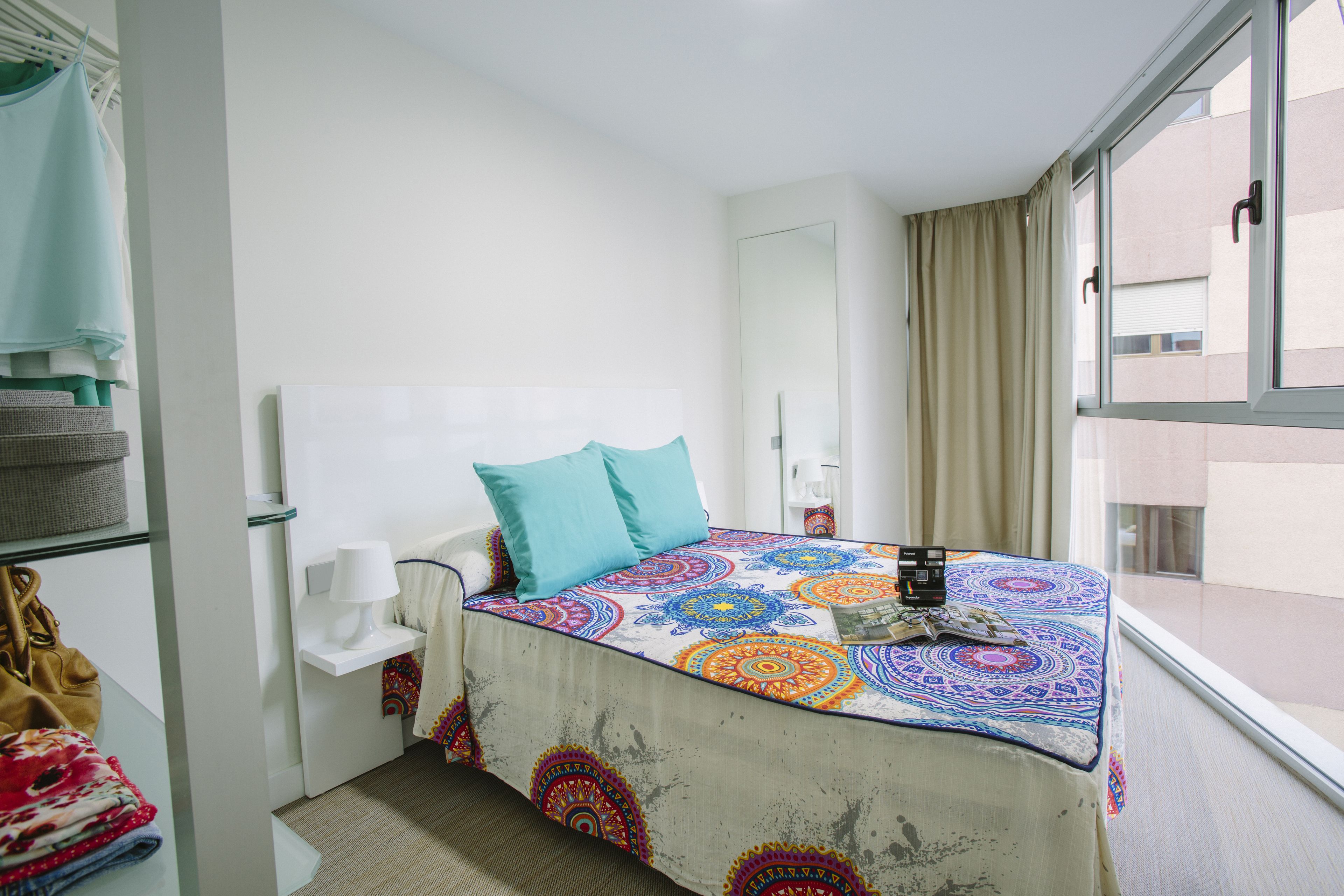 Habitación de hotel con cama, mesita de noche con lámpara, ventana con cortinas y vistas al exterior.