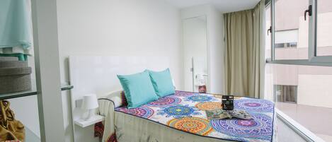 Habitación de hotel con cama, mesita de noche con lámpara, ventana con cortinas y vistas al exterior.