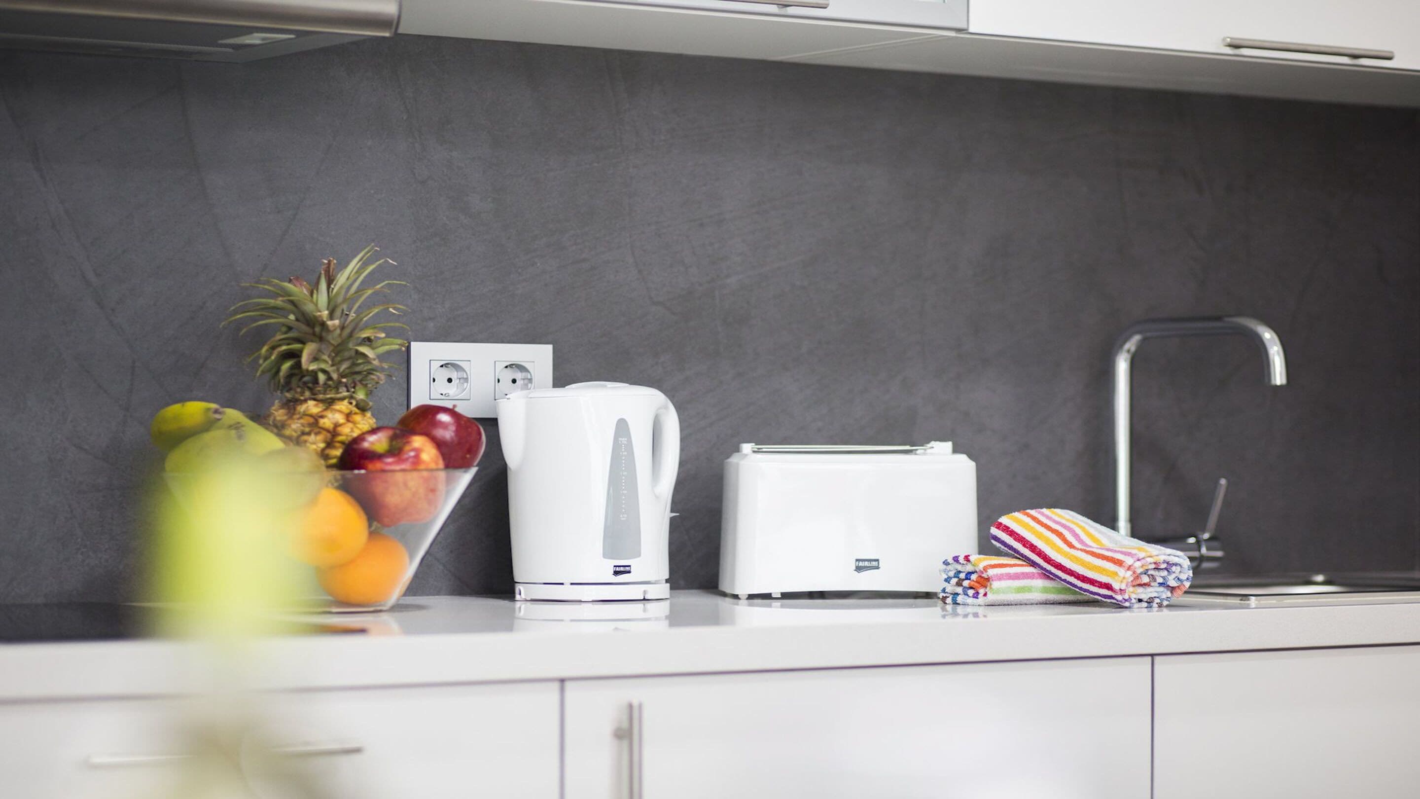 A modern kitchen with a white countertop, a kettle, a toaster, and a fruit bowl containing bananas, apples, and oranges.